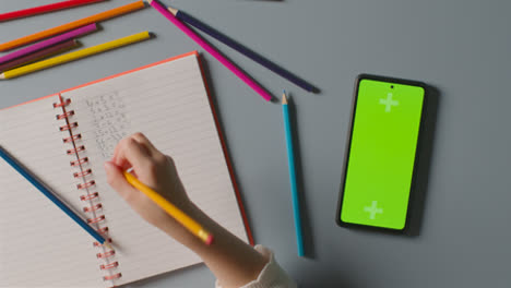 Overhead-Shot-Of-Child-With-Green-Screen-Mobile-Phone-Doing-School-Maths-Homework-In-Book-1